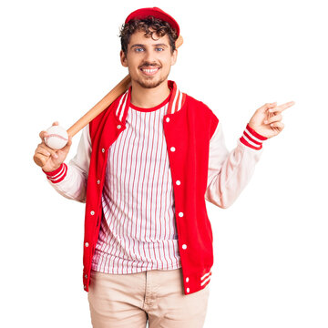 Young handsome man with curly hair playing baseball holding bat and ball smiling happy pointing with hand and finger to the side
