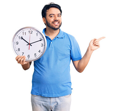 Young hispanic man holding big clock smiling happy pointing with hand and finger to the side