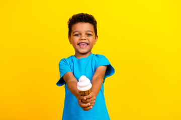 Portrait of adorable little schoolboy with afro hair wear blue t-shirt give you yummy ice cream isolated on vivid yellow background