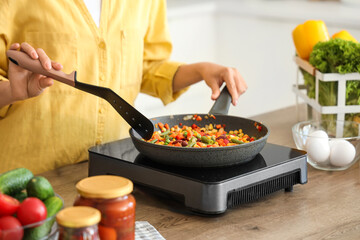 Young Asian woman cooking different vegetables in frying pan on portable electric stove at kitchen