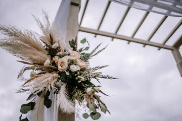 Close-up of a floral arrangement on a wedding arch, with a mix of pampas grass, roses, and greenery against a white structure and sky background.