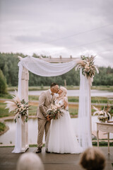 Valmiera, Latvia - Augist 13, 2023 - Bride and groom kissing under a wooden arch with floral...