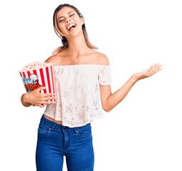 Young beautiful woman holding popcorn celebrating victory with happy smile and winner expression...