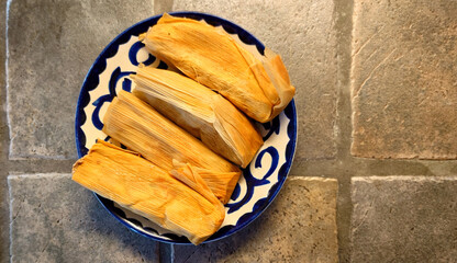 four pork tamales still in the husk on a painted blue and white plate on a porcelain tiled countertop  - 775254804