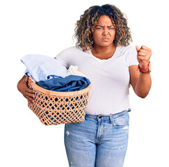 Young african american plus size woman holding laundry basket annoyed and frustrated shouting with...