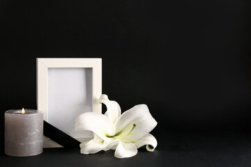 Beautiful lily flower with blank funeral frame and burning candle on dark background