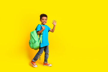 Full size photo of positive little schoolboy wear casual blue t-shirt hold rucksack waving palm say hi isolated on vivid yellow background © deagreez