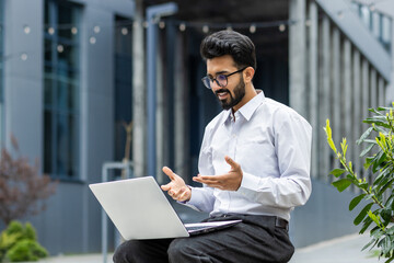 A young Indian male office worker is having a video call, sitting on a bench near a building and holding a laptop on his lap.