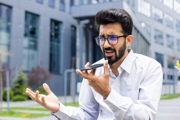 Worried and upset young Indian man in white shirt and glasses sitting near working office and talking on the phone through loudspeaker ,and recording voice message.