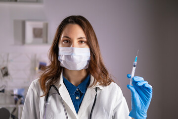 A young adult female doctor in a medical mask and white coat holds a syringe in her hands and looks at the camera