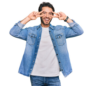 Young hispanic man wearing casual clothes doing peace symbol with fingers over face, smiling cheerful showing victory