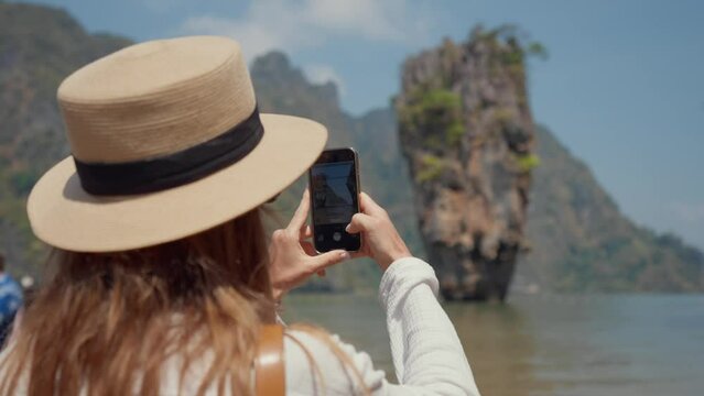 Woman travels on island Khao Phing Kan. Young female tourist sits on a boat and films take a photo of beautiful mountain landscape with a rock in the sea on smartphone. Travel, journey concept.