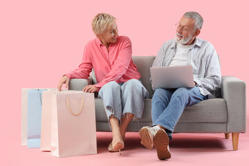 Mature couple with laptop and shopping bags sitting on sofa against pink background