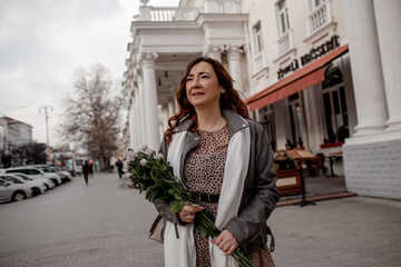 A beautiful middle-aged woman keeps a bouquet of beautiful flowers, purple roses. The girl walks around city and enjoys the cloudy wather