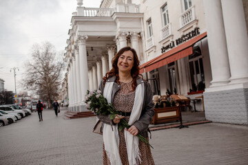 A beautiful middle-aged woman keeps a bouquet of beautiful flowers, purple roses. The girl walks around city and enjoys the cloudy wather