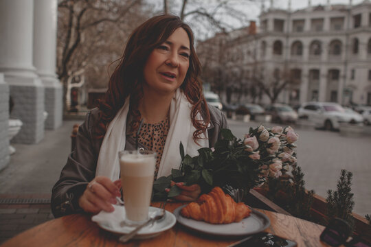 A charmig middle-aged woman drinks latte and eats  croissant in a french cafe on a city street. A woman with flowers is enjoing a wonderful and tasty breakfast