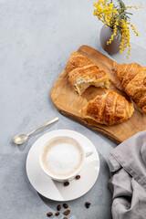 A cup of aromatic cappuccino coffee and fresh croissants on a cutting board on a blue background with coffee beans and flowers.