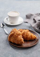 A cup of aromatic cappuccino coffee and fresh croissants in a wooden plate on a blue background with morning light.