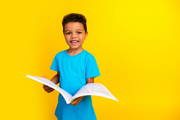 Portrait of good mood smart schoolboy with afro hair wear blue stylish t-shirt hold book learn lessons isolated on vivid yellow background
