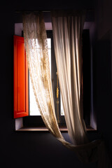 La luz del atardecer entra por una ventana cerrada con porticones naranjas a través de las cortinas en un casa vieja oscura.