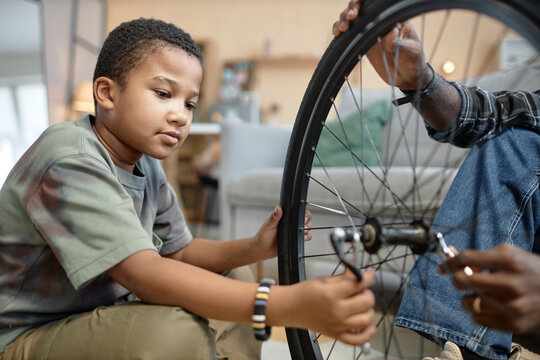 Side view portrait of young African American boy repairing bicycle tire with dad sitting on floor at home