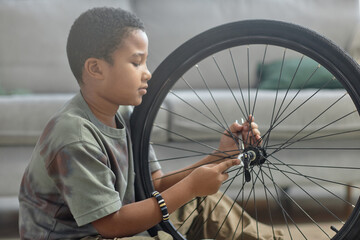 Side view portrait of African American young boy repairing bicycle wheel and checking tires, copy...