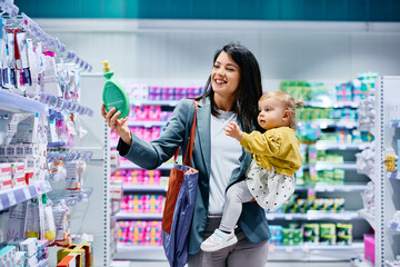 Happy mother choosing baby shampoo while shopping with her daughter in supermarket.