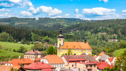 The Church of Saint Bartholomew in Pecka towers over quaint houses amidst lush green hills under a clear blue sky. Czechia - 775336848