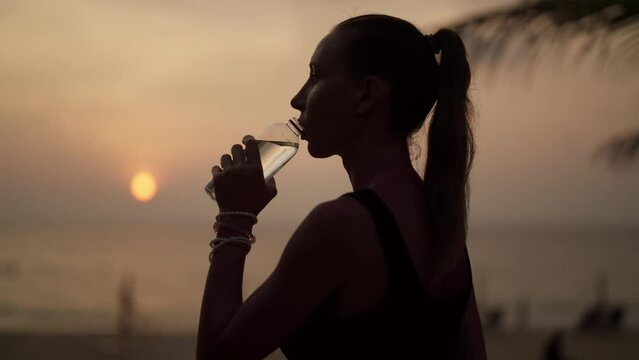 Woman drinking water after training. Fitness thirsty girl drinks water during workout pause, after a grueling run of a long sea ocean beach at sunset. Endurance sport, fitness, morning run concept.