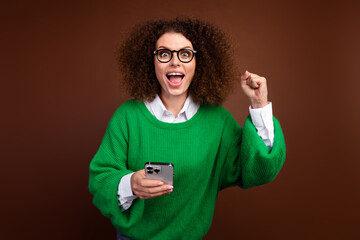 Portrait of attractive crazy lady hold smart phone raise fist shout yes hooray isolated on brown...