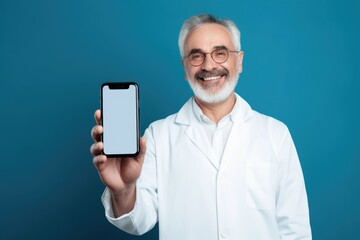 Happy senior male doctor showing a blank smartphone screen with copy space, on a blue background. Smiling Doctor Presenting Smartphone Screen