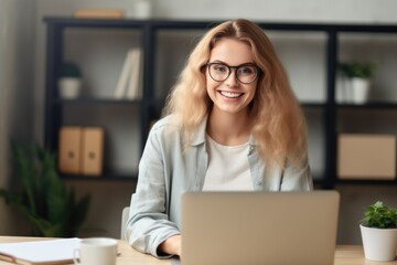 A cheerful young woman with glasses works on her laptop in a well-organized office space, displaying productivity. Smiling Woman Working on Laptop at Desk