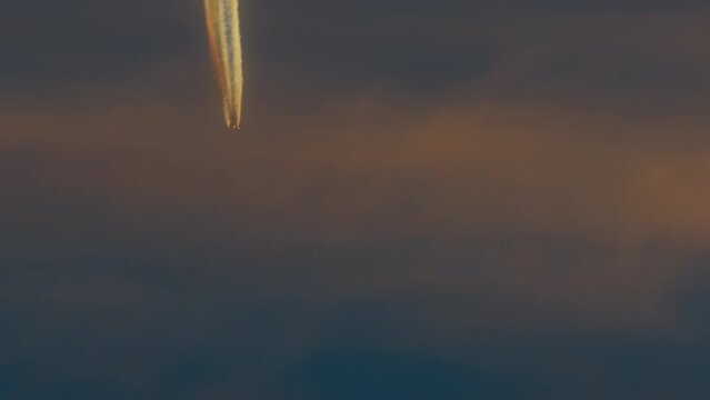 An airplane is seen flying at a high altitude in the sky during sunset, with distinct contrails against the orange-tinged clouds