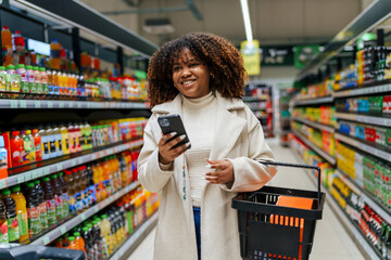 A curly hair Black woman with shopping basket using phone while buying and picking groceries in...