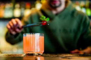 A bartender garnishes a pink cocktail with a cherry in a modern bar setting. The drink, prominently displayed in the foreground, is a vibrant pink color.