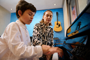 Female pianist, music teacher teaching piano and music lesson to a teenage boy in white casual...