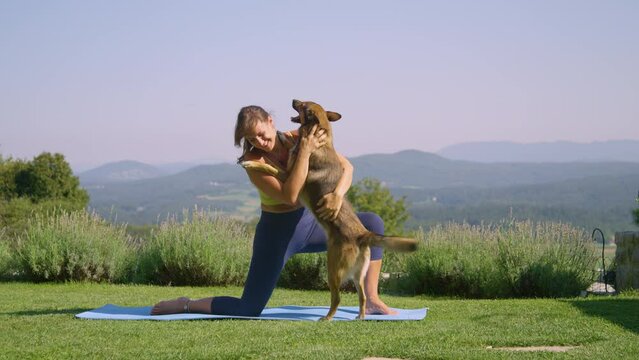 Woman practices yoga in garden when her playful and excited dog jumps at her. Lady tries to relax and strengthen her body through exercise while her silly and naughty young doggo just wants to play.