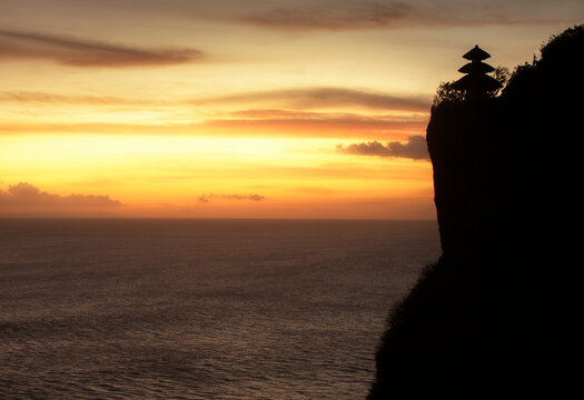 The silhouette of a tiered temple on a clifftop against the backdrop of a vibrant sunset sky over the tranquil ocean
