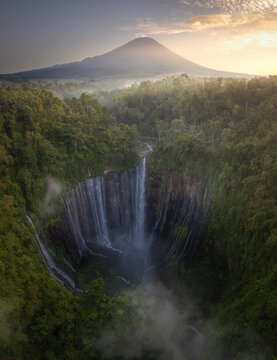 The first light of dawn illuminates a misty, majestic waterfall with a towering volcano in the distance, amidst the tropical forest of Indonesia
