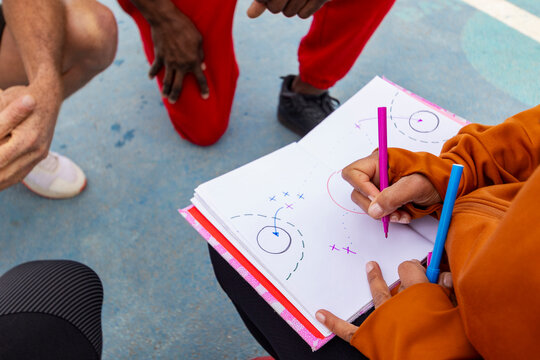 Close-up view of a coach's hands drawing a play on a basketball strategy board during a streetball session with players huddled around