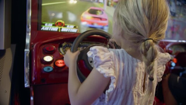 Blonde child in a ponytail playing a car racing video game, hands on the wheel at an arcade