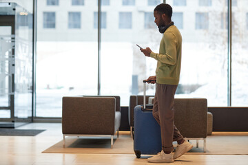 Side view full length portrait of African American man with luggage walking across hotel lobby and using smartphone calling taxi app