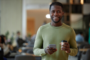 Waist up portrait of modern Black man holding smartphone and coffee cup smiling at camera in office...