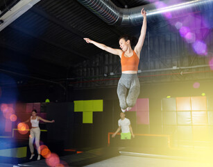 Positive middle-aged woman in sporty clothes jumping vertical high in modern trampoline hall