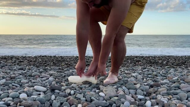 Video 4K. White adult bald man with hairy bare legs in yellow shorts puts on protective transparent shoes on pebble beach against backdrop of sunset, setting sun and sea waves, ocean. Travel, tourism