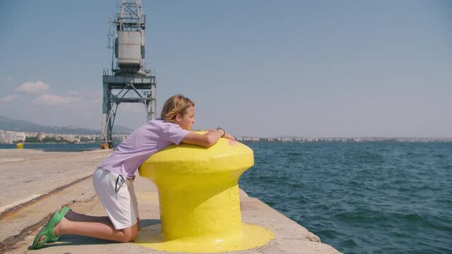 Boy in a purple t-shirt and white shorts leans on a yellow harbor bollard, looking out at the sea with a large crane in the background