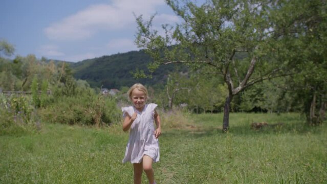 A girl in a white striped dress is captured in slow motion as she runs joyfully across a meadow, with a backdrop of trees, greenery, and hills