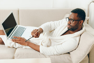 Smiling African American Man in Bathrobe Working on Laptop on Sofa in the Morning A young African...
