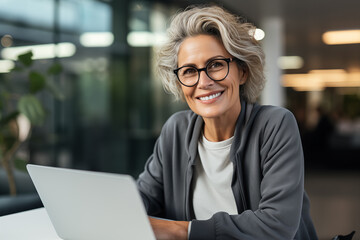 A mature businesswoman in her 50s with silver hair smiles while using her laptop in a casual cafe...