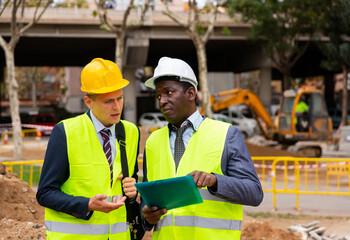 Two men workers working at the laying paving slabs facility discuss the project, holding important...